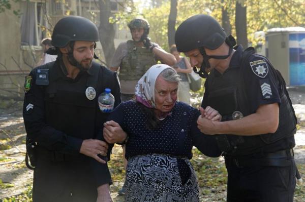 Rescue workers escorting an elderly woman from her flat after it was hit by a Russian bomb. She is wearing black and has a white scarf tied around her head.